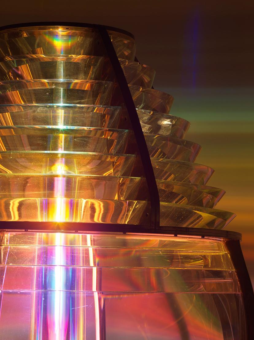 A detail view of stacked, curved glass lenses, with horizontal bands of gold, pink and green, refracting colour into vivid horizontal rays.