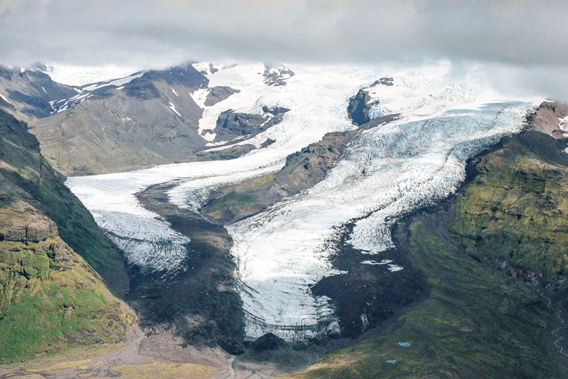 An aerial view of a broad, branching glacier descending through a mountain valley, with low clouds hanging over its upper snowfields and rocks protruding from parts of its icy surface.