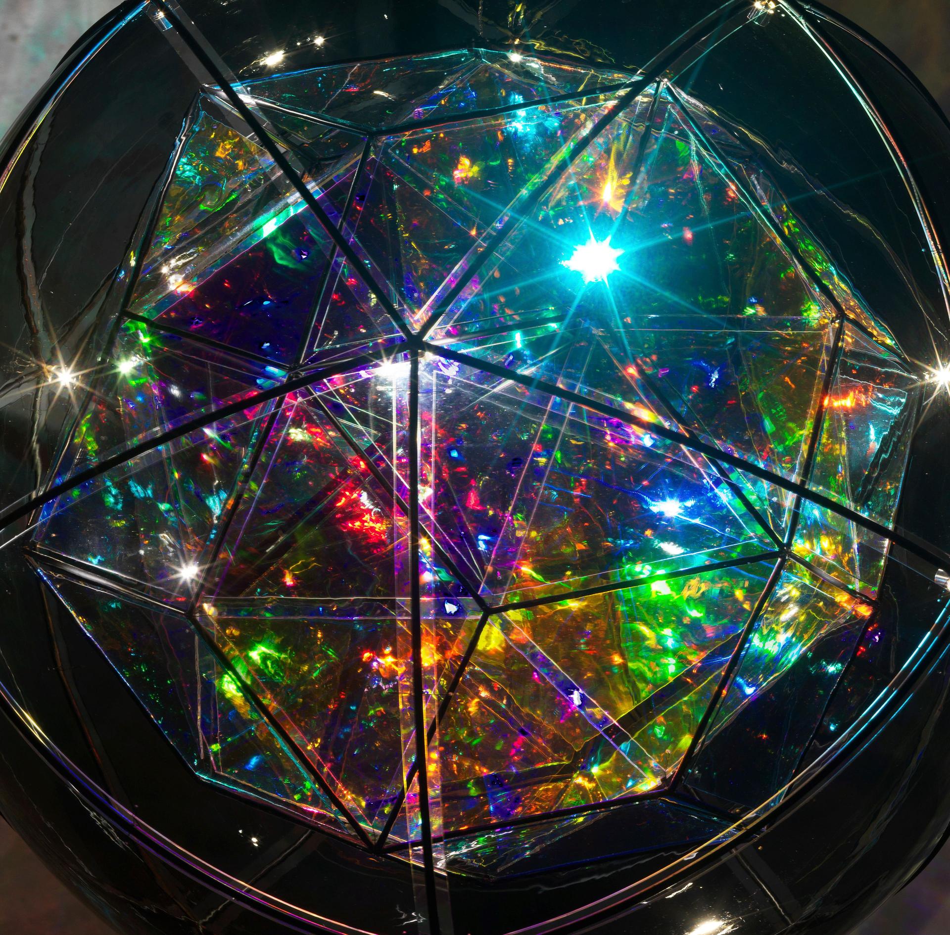 The multi-faceted glass form shimmers with intense light, each pane reflecting different saturated colours within the mirrored interior, creating a dense field of rainbow colours.