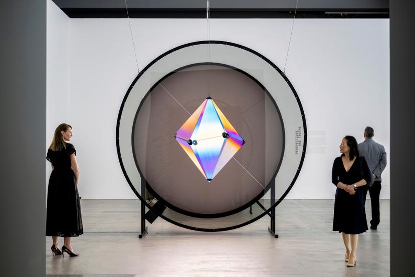 A large circular frame holds a faceted geometric form glowing from within, its translucent panels shifting between blue, purple, yellow and pink around dark circular connectors, with three visitors standing nearby.