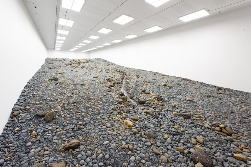 A sloping terrain of grey and tan stones filling a long, rectangular, white-walled gallery, with a narrow stream of water winding down the centre beneath bright ceiling lights.