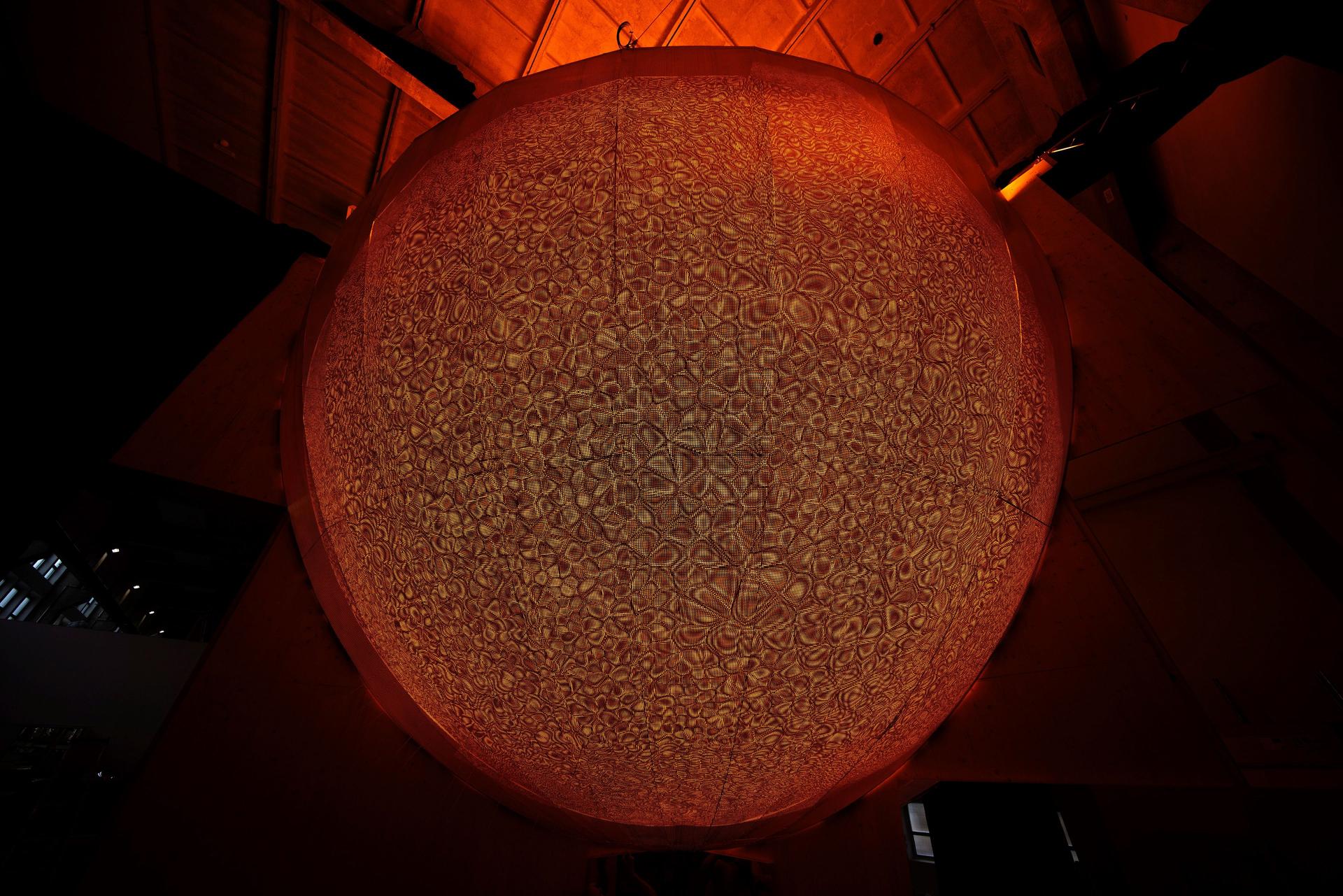 A large glowing sphere dominates a dark industrial space, its surface densely patterned with small, curved shapes.