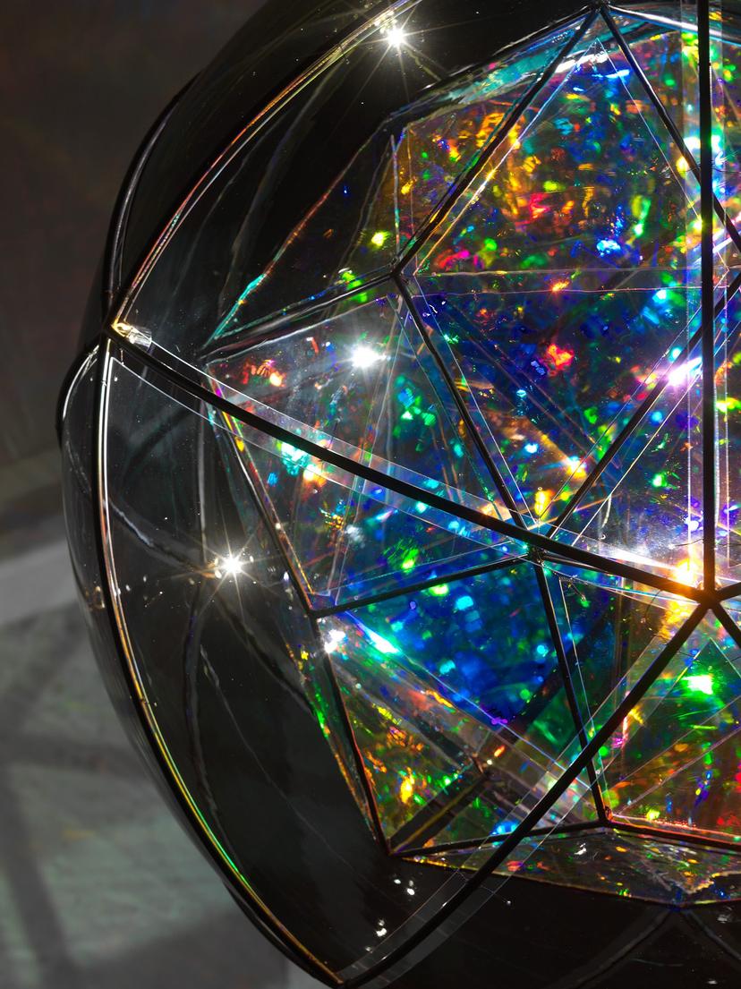 A close-up of a prismatic, spherical sculpture’s inner shell, showing overlapping triangular panes that refract vivid hues of green, blue, yellow, and red, framed by the ribs of a black metallic frame.