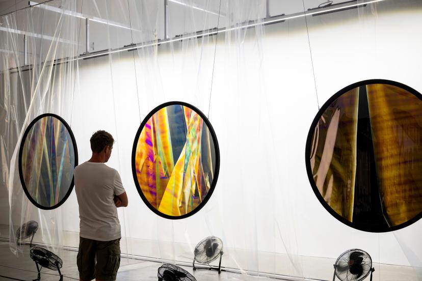 A person stands in front of three suspended circular polarising filters, each showing the translucent curtain behind them in shifting tones of yellow, blue, brown and iridescent multicolour, framed by hanging transparent sheets.