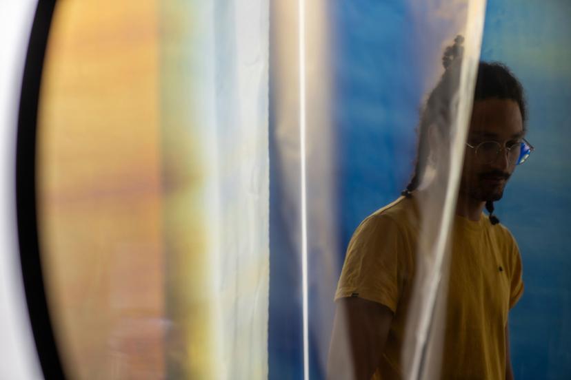 A partially obscured person in a yellow shirt appears behind translucent vertical panels, with soft reflections and blurred blue and yellow tones across the surface.