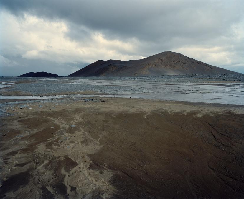 A wide, muddy wetland plain with branching, water-filled channels weave toward distant low hills, with pale grey patches of ground and a cloudy sky above.