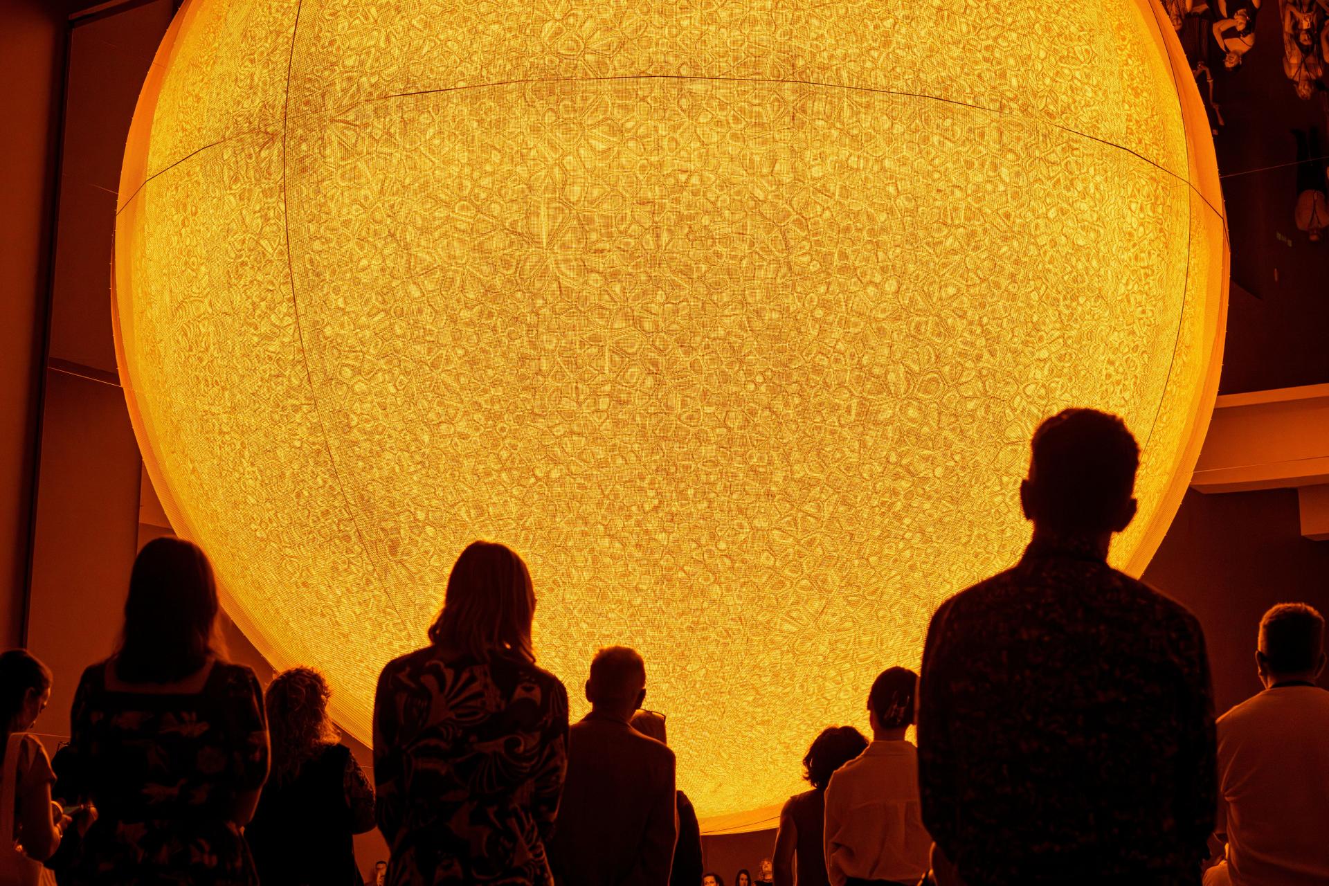 Silhouetted visitors gather beneath a vivid yellow spherical artwork that fills the upper frame, its surface marked by tightly clustered curved patterns activated by the motion of viewers.