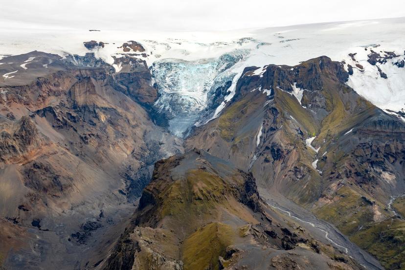 An aerial view of a glacier with a visibly truncated lower section, set between steep brown and green mountains.