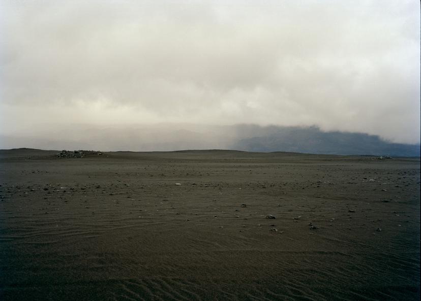 A wide expanse of dark, granular ground scattered with small rocks extends toward low, cloud-covered hills beneath a dense grey sky.