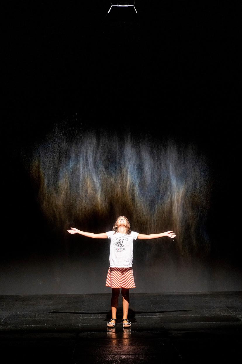 A child stands on a dark floor with arms outstretched, looking up towards a vertical sheet of fine mist illuminated with soft blue, amber and violet tones beneath a small overhead light.