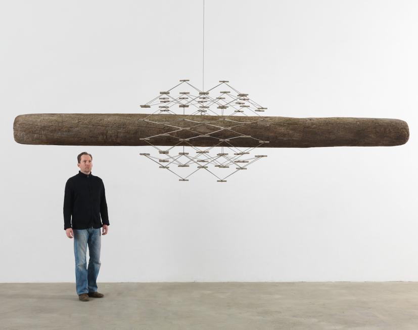 A man stands inside a white-walled gallery beside a long, suspended log that is intersected at its centre by a complex, geometric, metal structure.