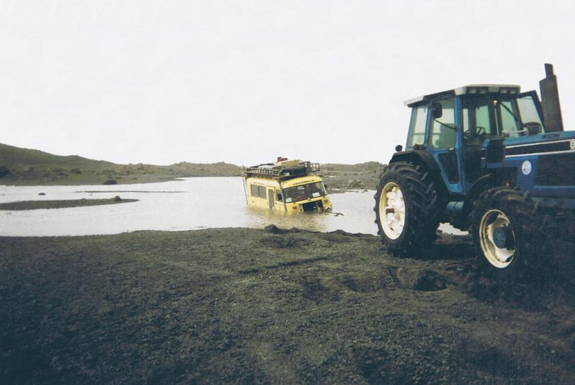 A yellow minibus sits partially submerged in shallow water while a blue tractor stands on a dark gravel bank in the foreground, with low hills in the distance.