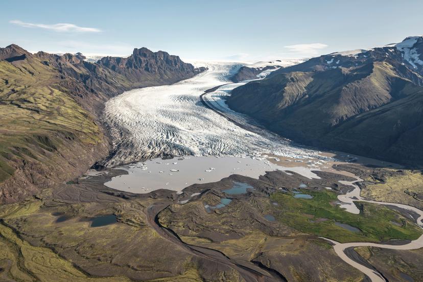 An aerial view of a glacier wide across its upper sections and ending abruptly in a pale meltwater lagoon, set within green moss-covered terrain and flanked by dark mountain ridges.
