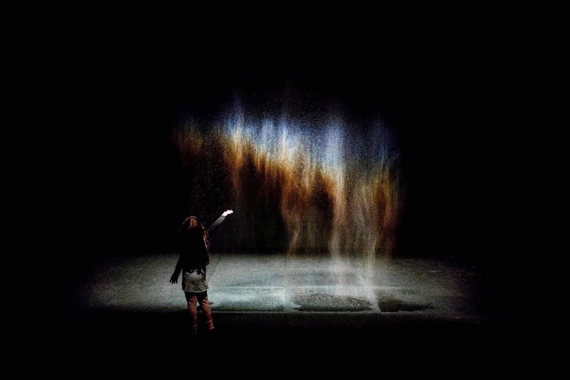 A young person stands before a sheer drift of mist, reaching towards the falling spray as blue, gold, violet and red hover around its droplets, creating vertical washes of colour in the surrounding darkness.