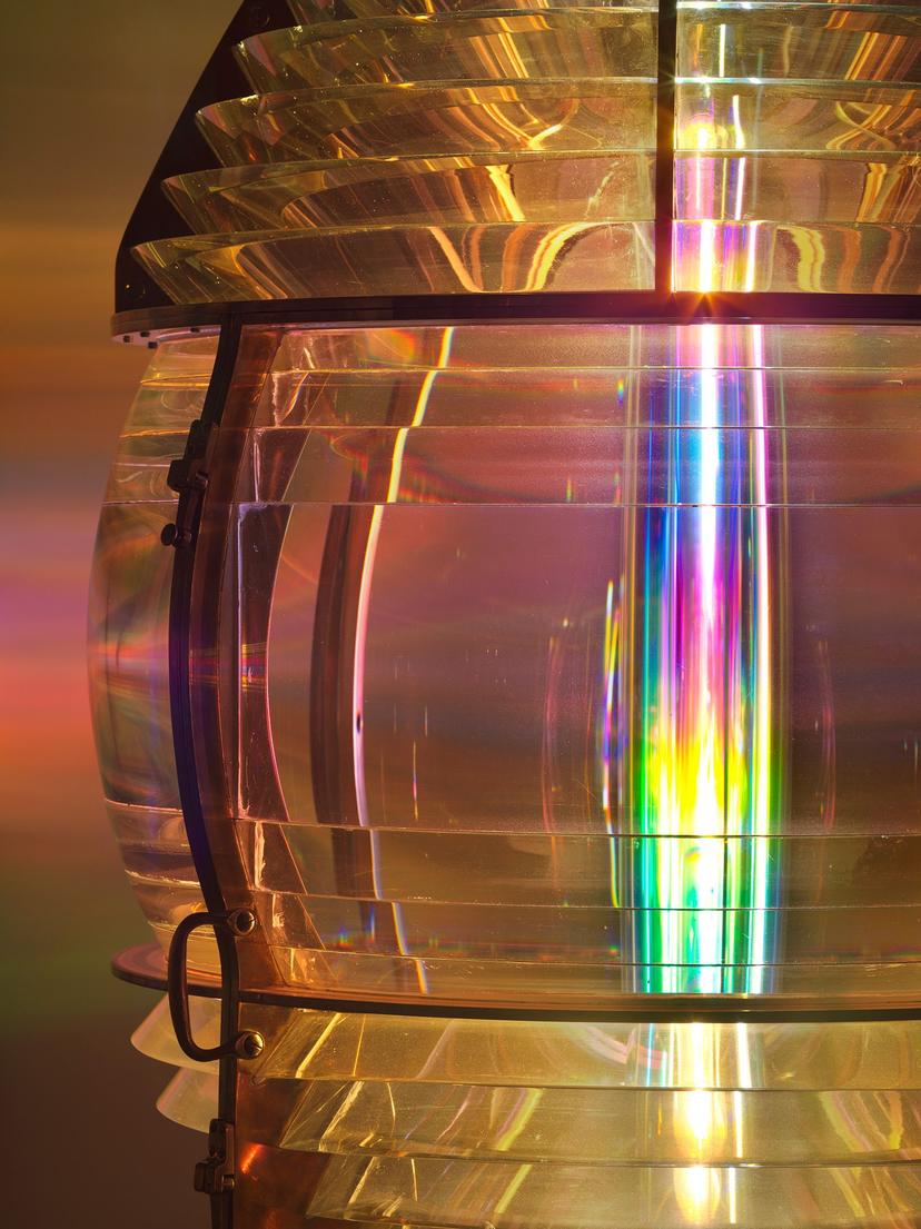 A side view of the illuminated lens revealing its brass fittings and clear glass structure. A vertical beam of multicoloured light runs through the centre, projecting a rainbow onto surrounding walls.