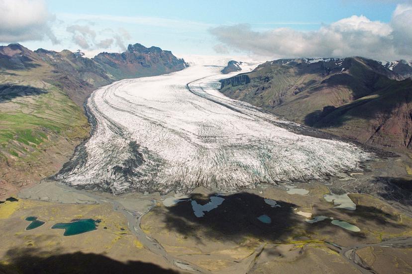 An aerial view of a broad glacier descending between two mountain ranges, its white surface marked with dark crevasses, ending in a flat valley dotted with pools and branching meltwater channels.