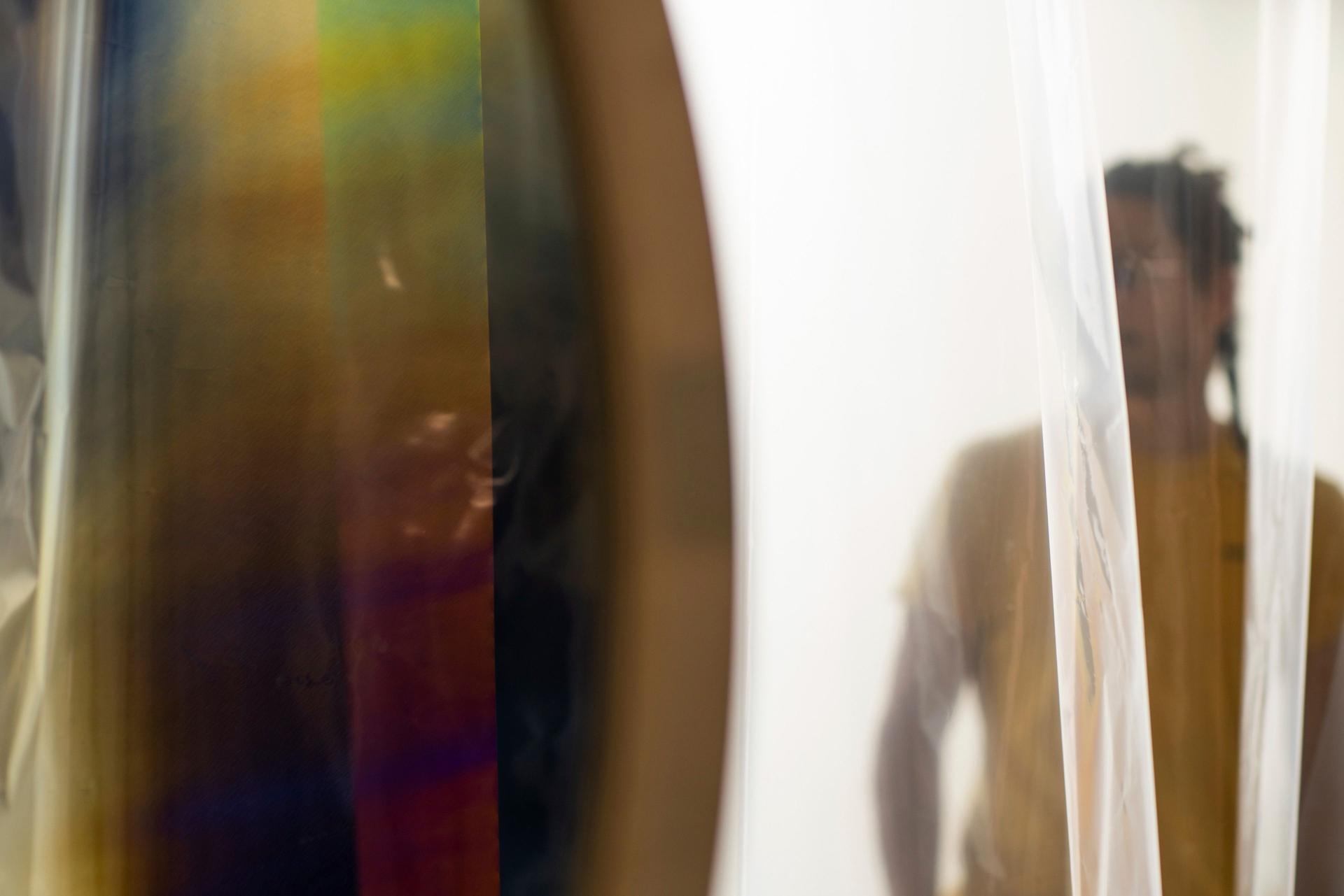 A blurred figure stands behind translucent hanging sheets, seen beside a large circular surface with soft, vertical bands of muted colour.