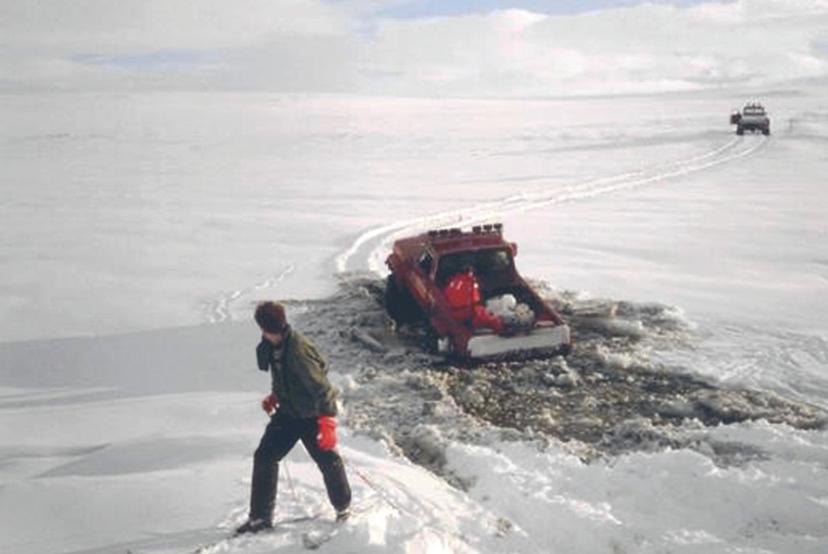 A red utility truck sits partially sunk in broken, slushy ice while a person walks nearby on firmer snow, with another vehicle visible in the distance across the snow-covered expanse.