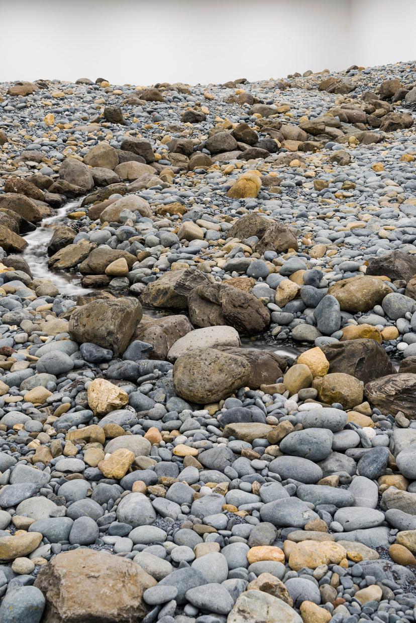 A gently curving stream of water winds through a wide expanse of grey and tan rocks and pebbles in a white-walled gallery.