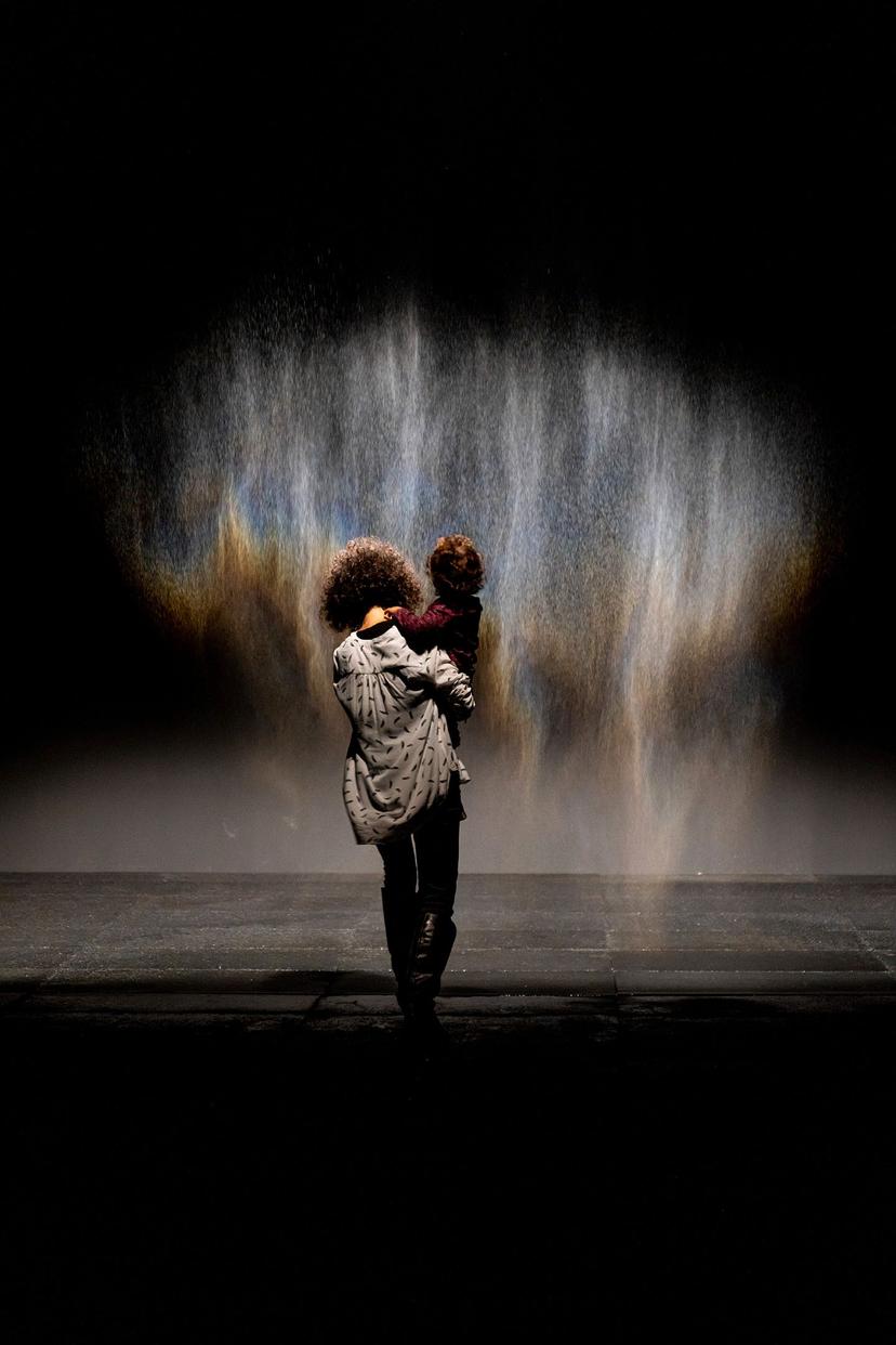 A person stands on a dark floor holding a child, both facing a fine, falling veil of mist alight with soft blue, amber and violet tones.