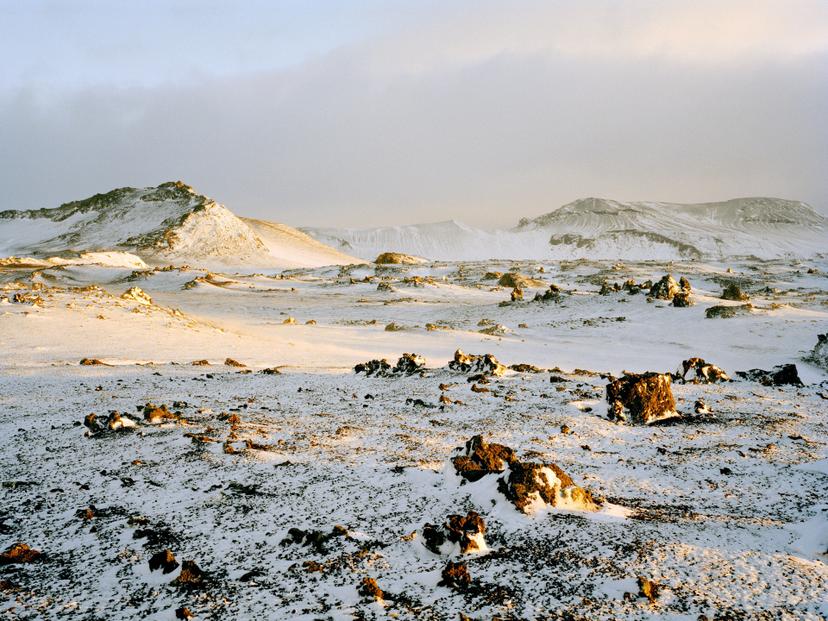 A snow-covered volcanic plain illuminated by warm light hitting uneven rock formations, with distant ridgelines visible through a faint mist.