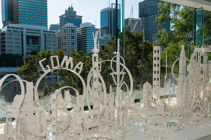 A dense arrangement of white plastic building-brick structures sits on a display table, forming tall, varied architectural shapes and circular frameworks. Among them is a large arched form spelling ‘GOMA’. Behind the installation, floor-to-ceiling windows reveal leafy trees, the river and numerous city buildings under bright daylight.