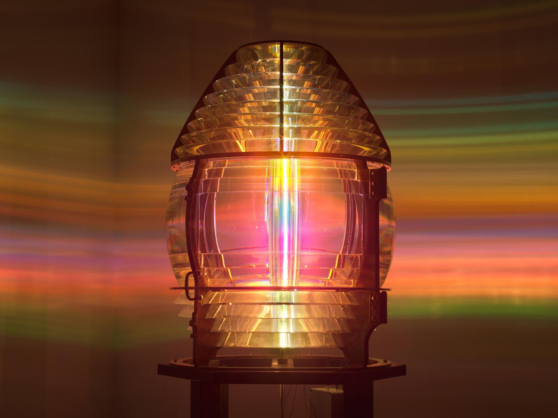 A glowing lens on a wooden plinth, casting circular rays of coloured light across the surrounding gallery walls. The projected light forms a radiant halo in the space.