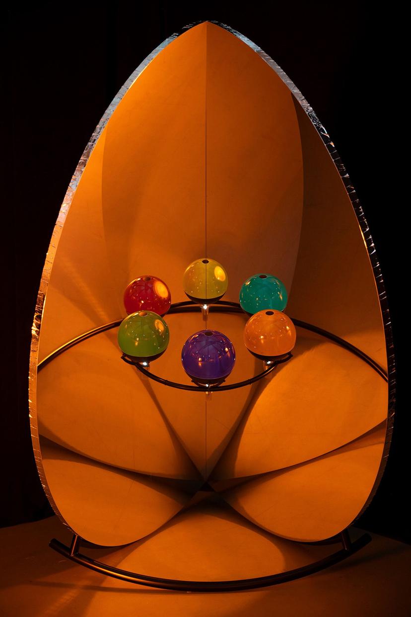A frontal view of six glossy coloured spheres mounted on a circular frame, set within a large petal-shaped surface that catches warm orange light and mirrors soft, curved patterns across its surface.