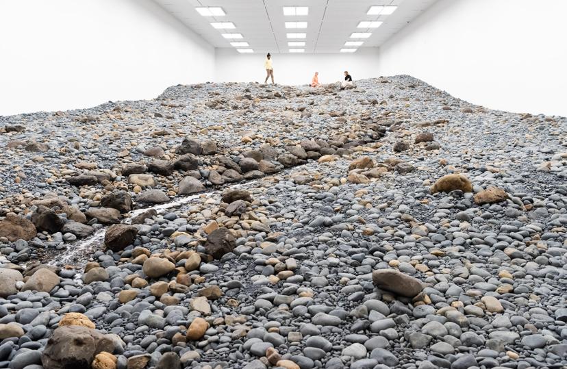 A wide view of a white-walled gallery filled with grey and tan rocks and a small winding stream, with three people visible in the distance under ceiling lights.