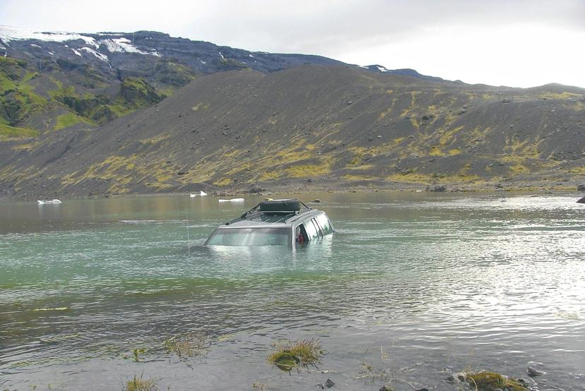 A four-wheel drive vehicle is partially submerged in a river below dark volcanic hills and green moss, with snow-covered peaks in the distance under a pale sky.