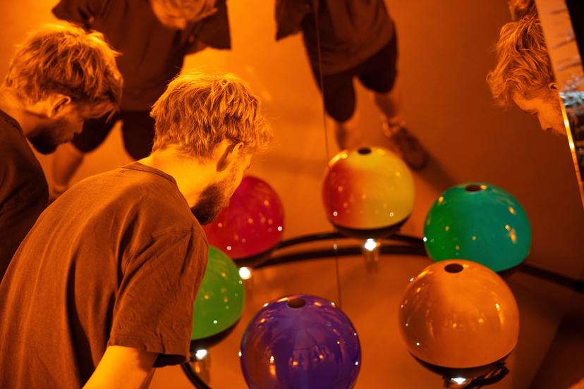 A close-up of a person leaning over a circular frame holding glossy coloured spheres, which appear blue, teal, green and amber, while multiple reflections of their figure appear in the tall mirror behind the arrangement.