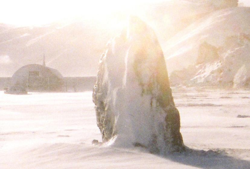 A backlit snowy scene with a vertical, ice-coated rock formation, its edges glowing in a bright haze, with a low dome-shaped structure visible in the background.