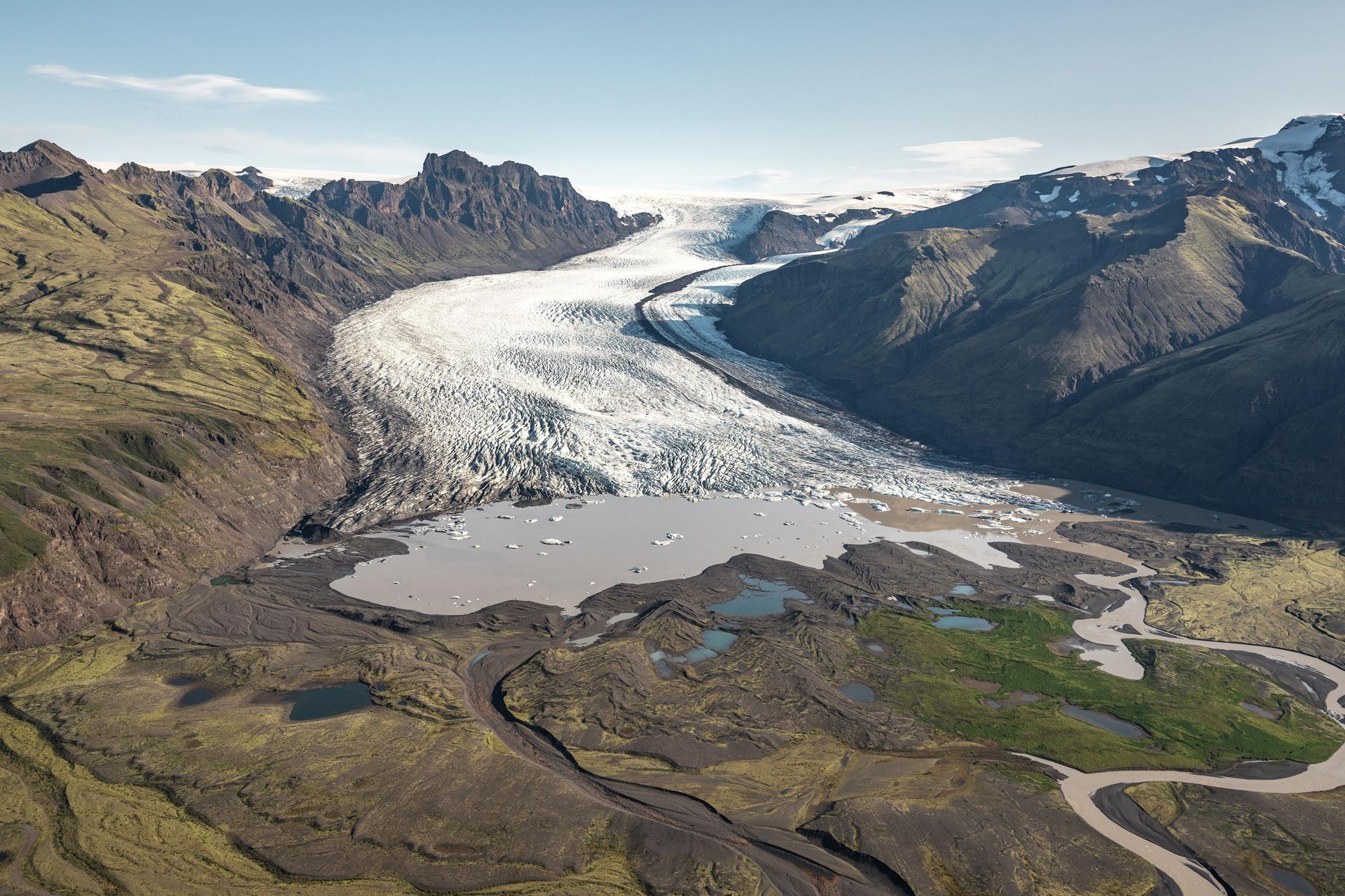 An aerial view of a glacier wide across its upper sections and ending abruptly in a pale meltwater lagoon, set within green moss-covered terrain and flanked by dark mountain ridges.