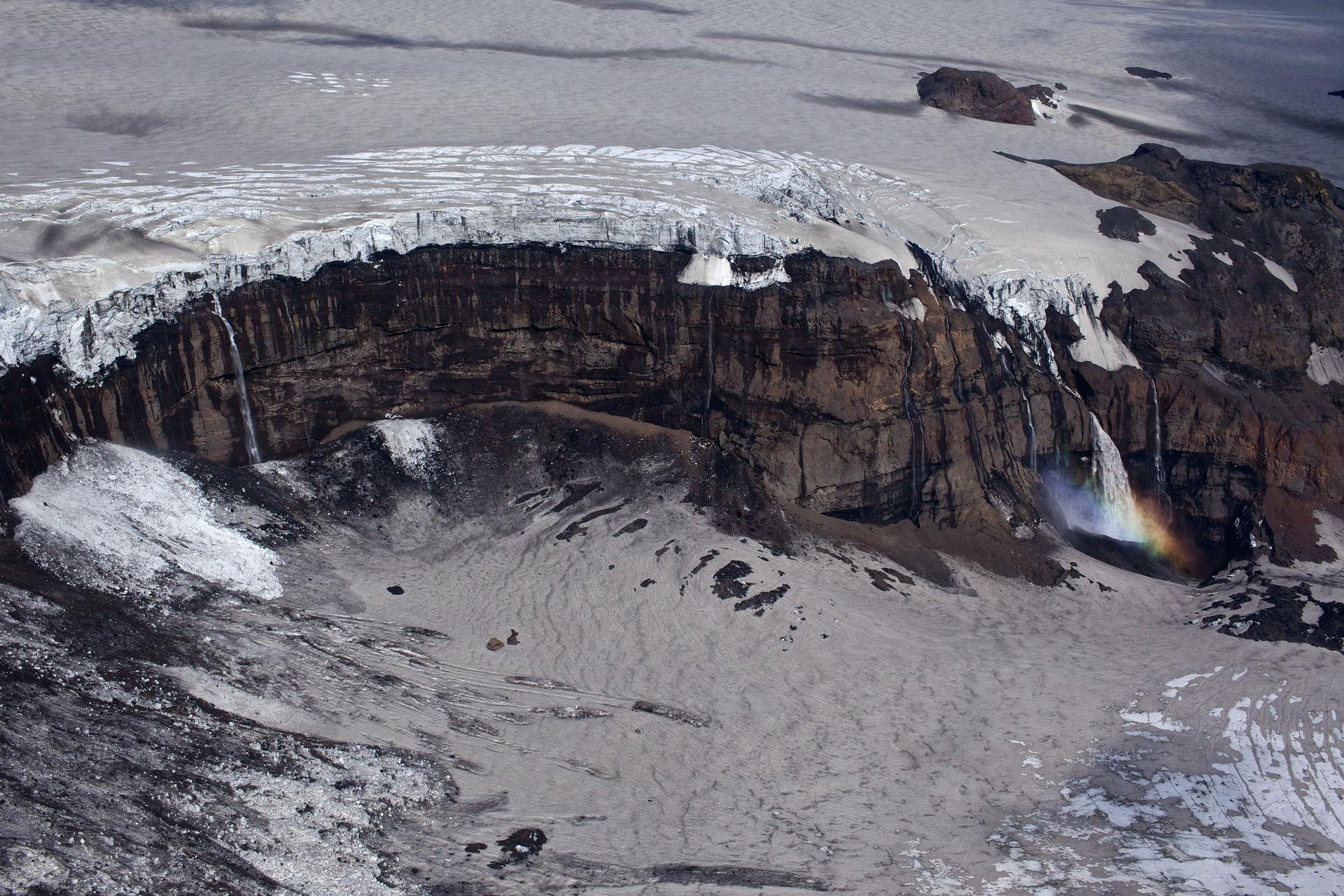 An aerial view of a glacier spilling over a dark volcanic escarpment, its meltwater creating waterfalls and a rainbow in a shroud of mist near the surface of the water.