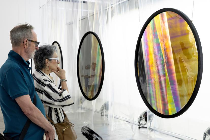 Two visitors stand beside a series of suspended circular polarising filters that display varied colour bands – yellow, blue, grey and multicoloured – visible through layers of overlapping translucent curtain material.