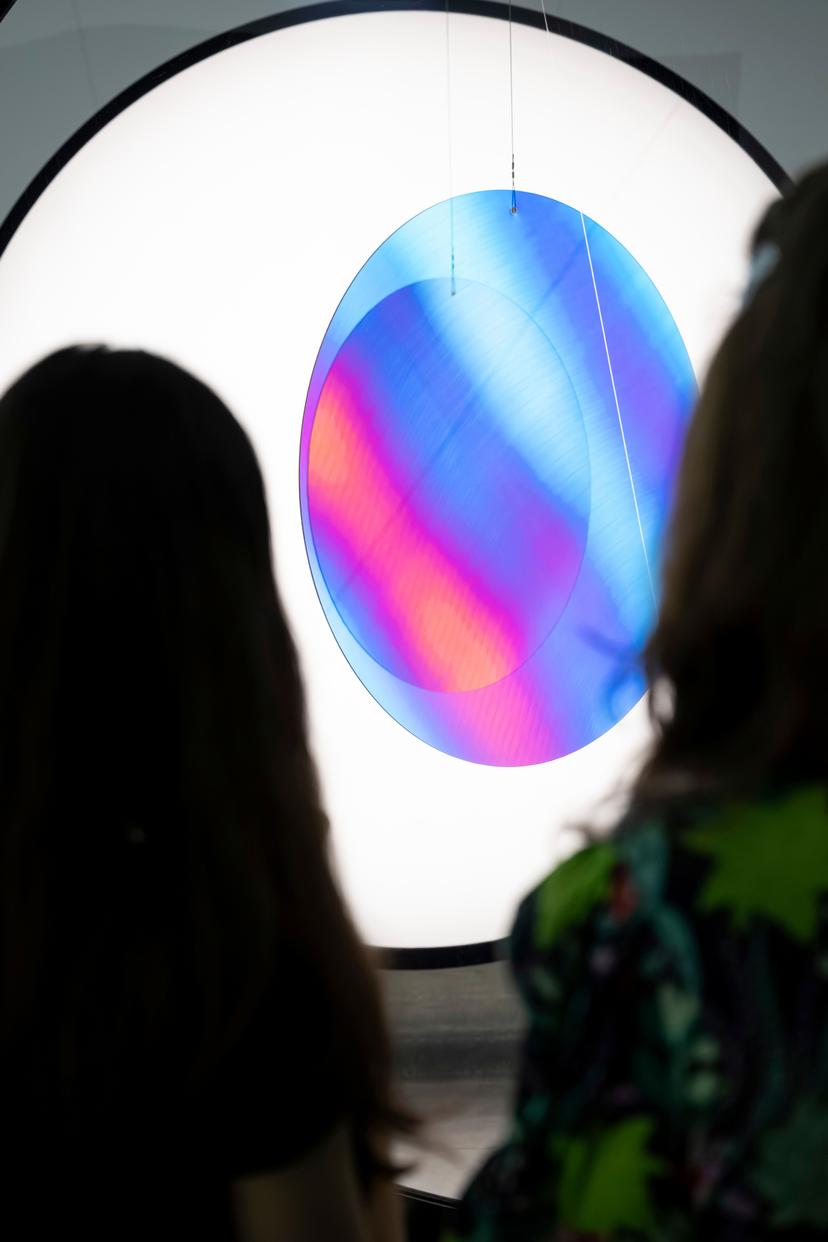 Two silhouetted visitors stand in front of a circular frame containing a luminous oval shape with shifting blue, purple and pink tones created by overlapping polarising layers.