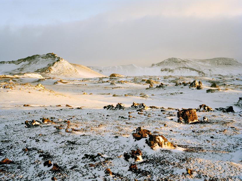 A snow-covered volcanic plain scattered with dark rock formations, illuminated by soft yellow light, with low ridgelines rising under a faintly golden sky.