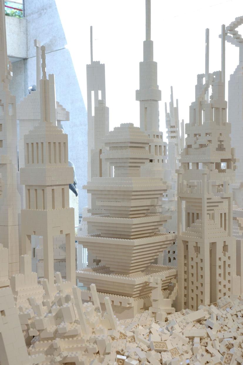 A dense cluster of white plastic building-brick structures with tall vertical towers, narrow window-like openings, stepped roofs, arched elements and zigzagging staircases. Loose bricks lie at the base, and the interconnected forms rise closely together.