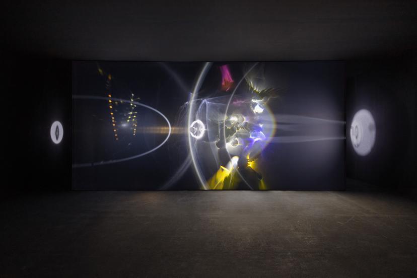 A person stands in a spacious dark gallery facing a tall white wall covered in sweeping bands and arcs of bright light and rainbow colours, including broad white curves and dispersing yellow and blue streaks that extend across the wall’s length.