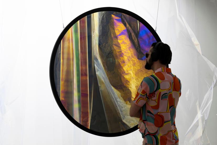 A close-up of a circular polarising filter reveals layered translucent sheets in bright yellow, blue, and dark iridescent colours, with a person nearby seen through the hanging transparent panels.