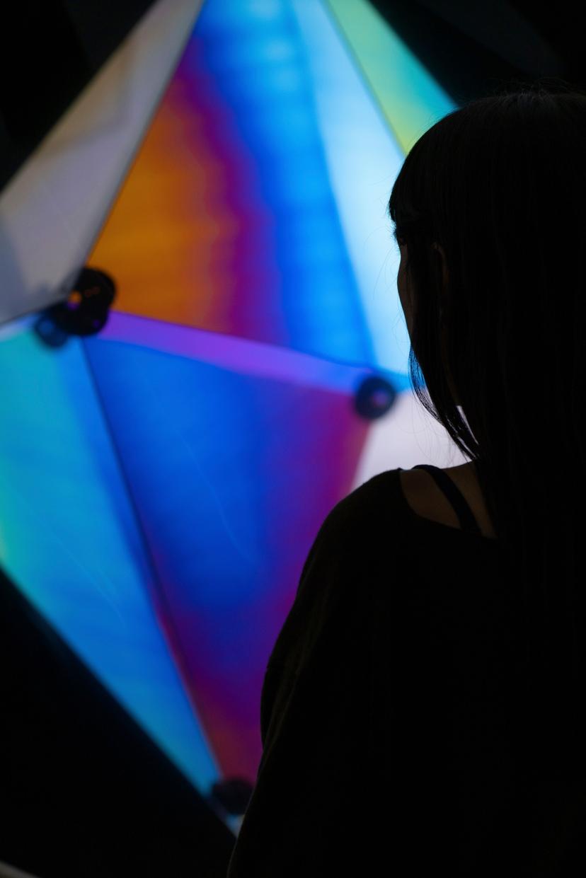 A person in partial silhouette stands before a glowing geometric form whose translucent panels blend blue, purple, yellow and orange tones around dark circular connectors.