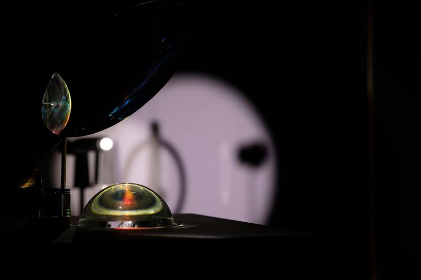 A transparent dome on a dark display surface contains coloured rings of light beneath a tilted reflective disc, set before a circular projection of soft white light.