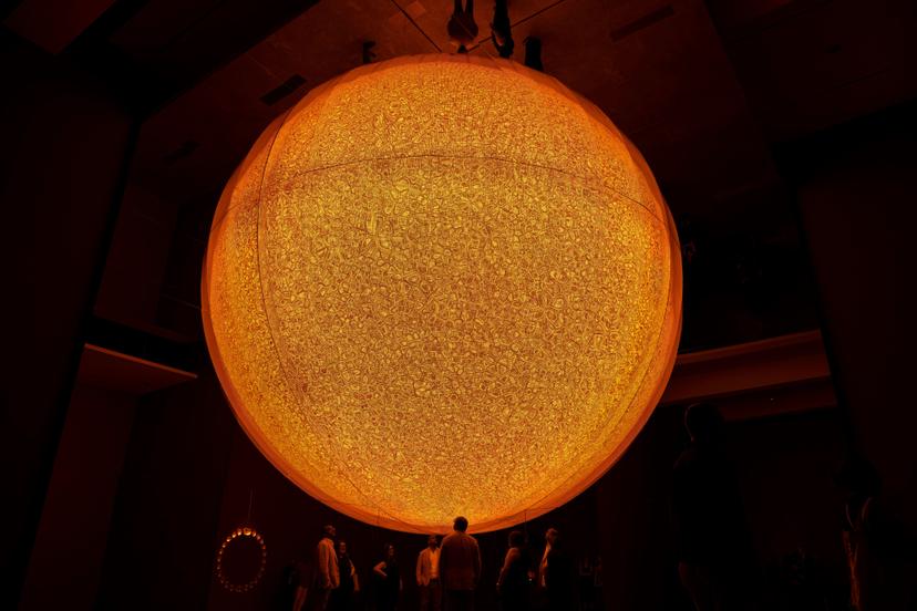 A large circular artwork glows with intense yellow light against a dark gallery ceiling, its textured surface composed of layered mesh and patterned fabric that produces a shifting moiré effect.