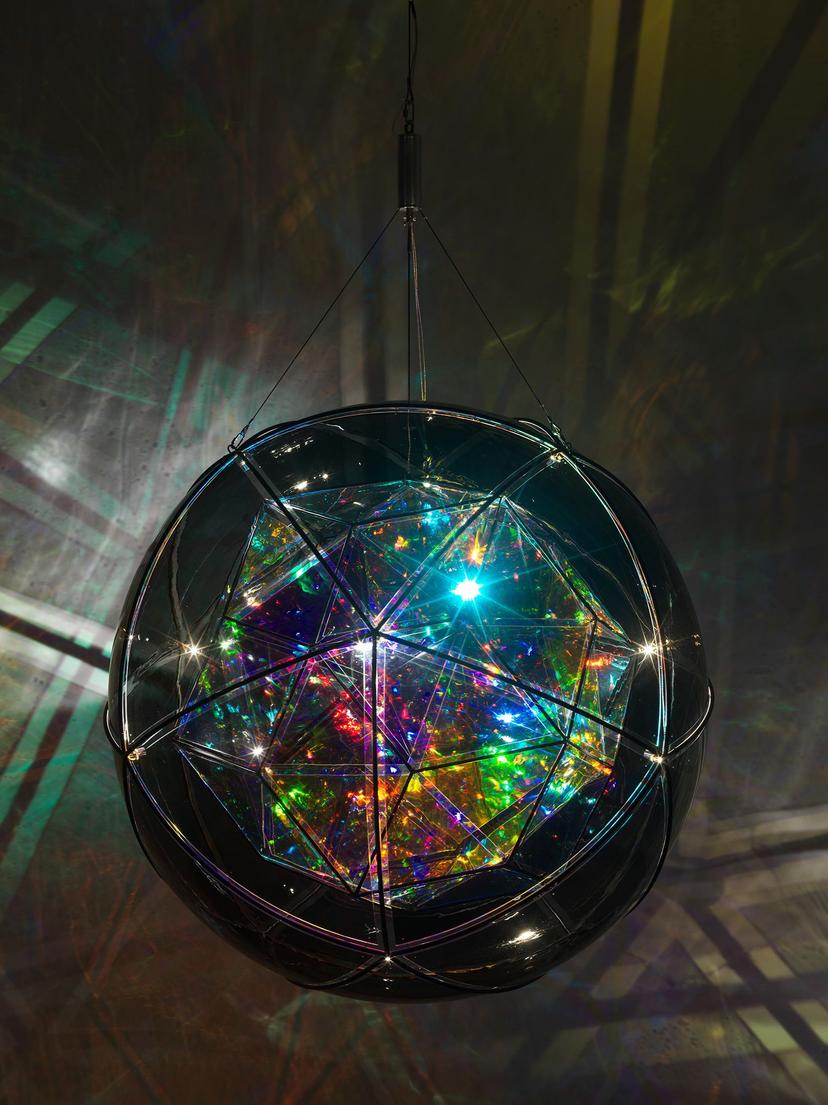 A large, illuminated geometric sculpture with a faceted, iridescent core hangs in a darkened space, projecting kaleidoscopic patterns of coloured light onto the surrounding walls.