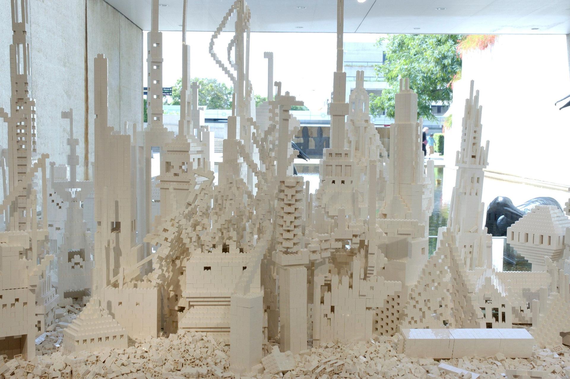 A dense grouping of white plastic building-brick structures forms tall, intricate towers, stacked platforms, arches and stepped shapes, rising from a layer of loose bricks. Behind the installation, large windows show a shallow water feature, part of a dark pelican sculpture, trees and people walking outside in daylight.