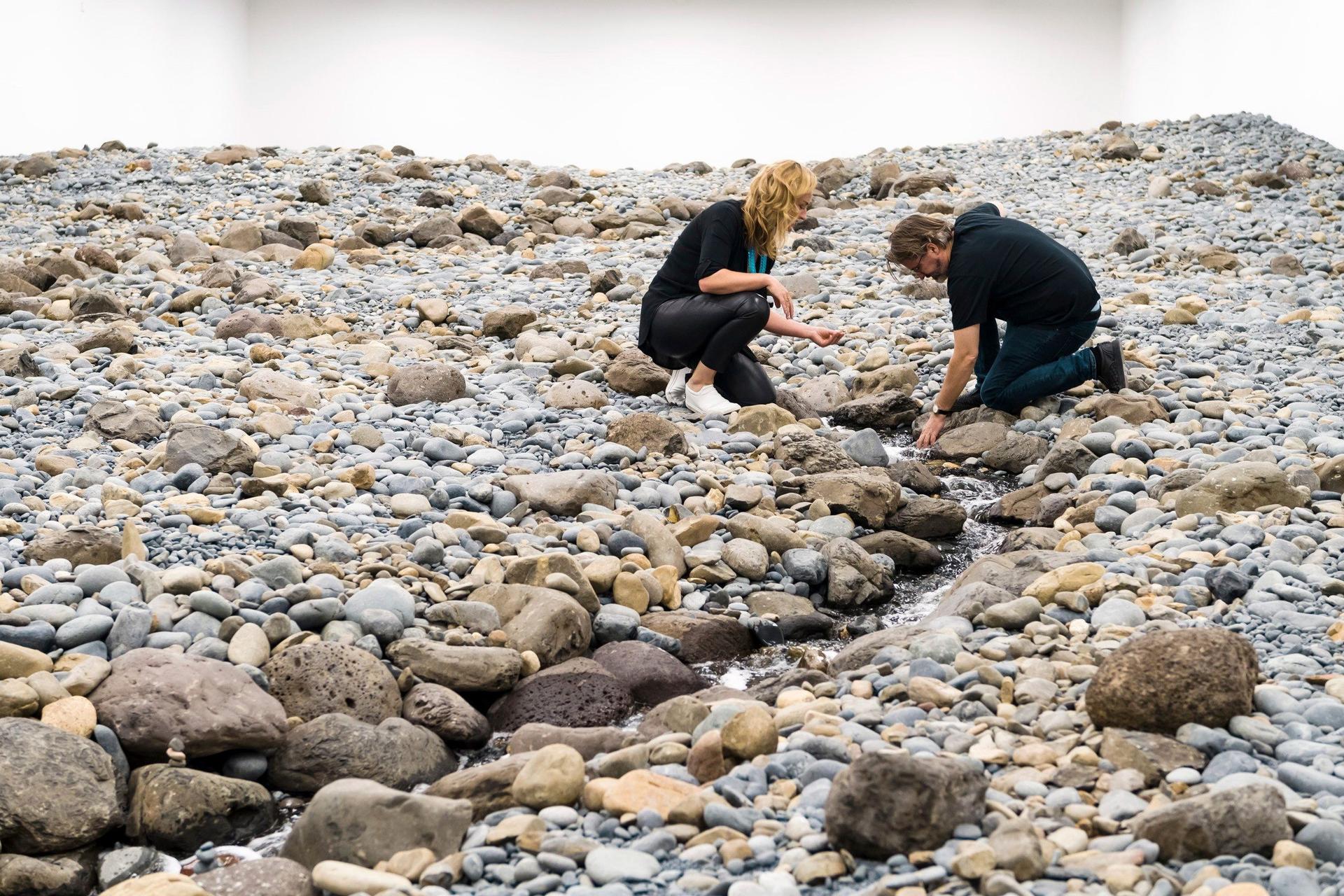 A person crouches on a rocky surface and extends a hand into a thin stream of running water that winds between grey and tan stones in a white-walled gallery.