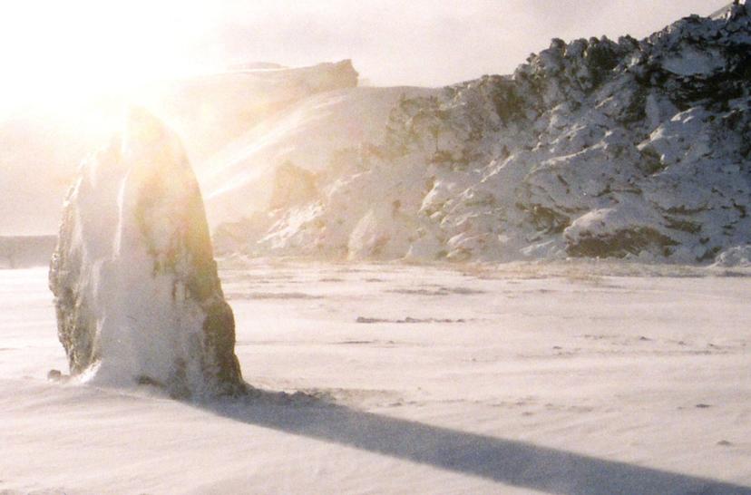Snowy terrain with a vertical, ice-covered rock in the foreground casting a long shadow, and bright backlight diffusing over the surrounding snow-covered hills.