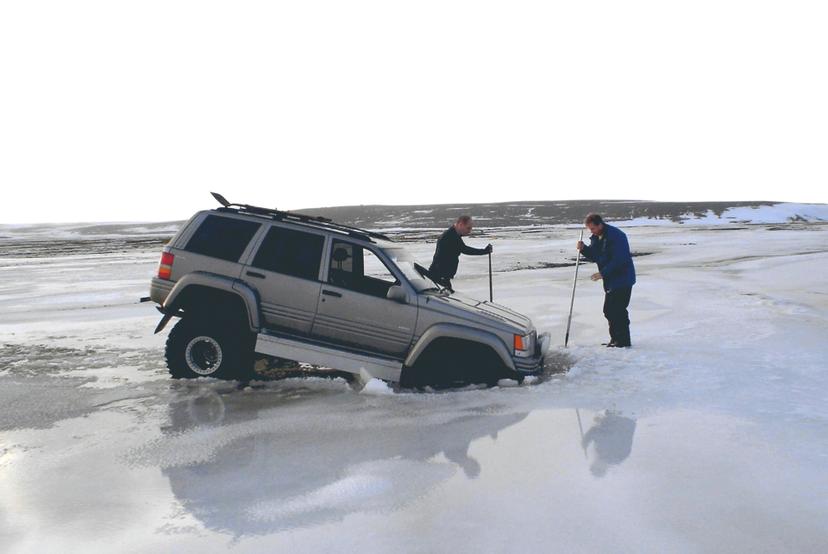 Two people stand beside a silver four-wheel drive vehicle whose front end is stuck in broken ice, using poles to probe the frozen surface in a flat, pale horizon.