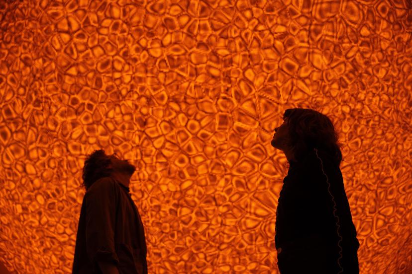 Two people stand in silhouette, looking up at a large, curved overhead surface that is glowing with bright orange light and covered in densely arranged clusters of small, curved forms.