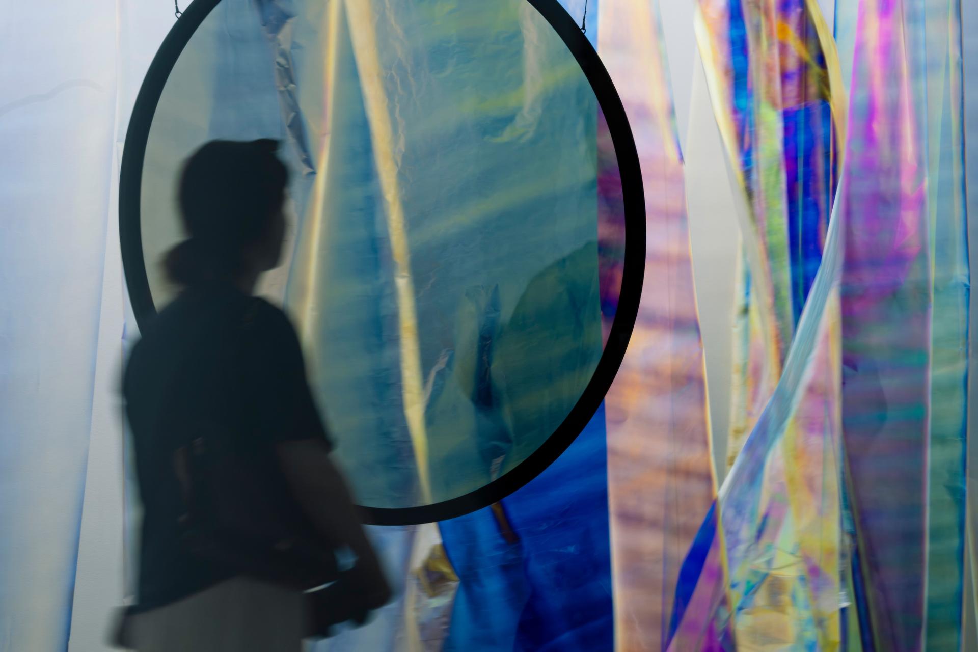 A silhouetted visitor stands before a large circular polarising filter that reveals layered translucent sheets in soft blue, green and yellow tones, with iridescent vertical strips in blue, pink and gold visible beside it.